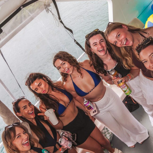 A group of smiling women in swimwear and summer outfits pose together on a boat, holding drinks. The background shows water and part of the boat's interior. They appear to be enjoying a celebration or party.