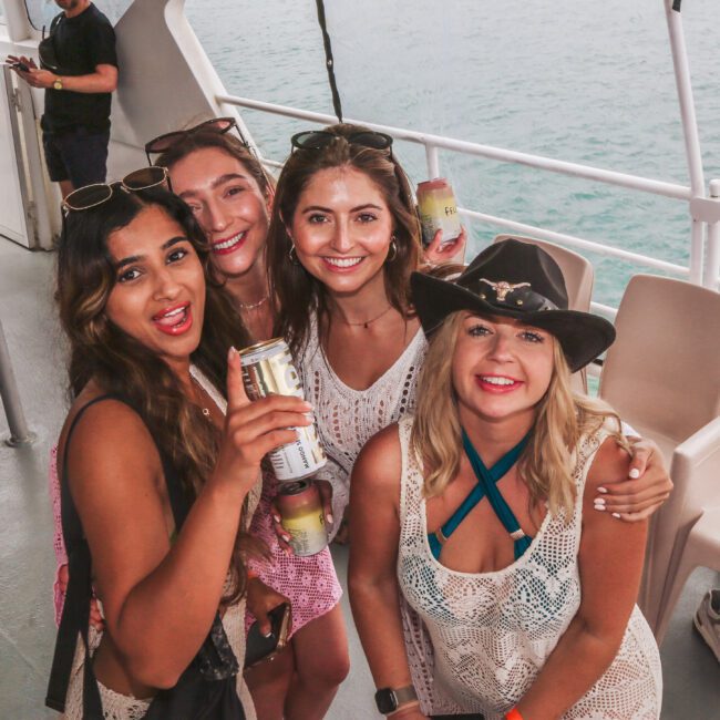 Four women smiling and holding drinks pose together on a boat deck with tan seats, with the ocean visible in the background. One woman wears a black cowboy hat; all are dressed in summer attire.