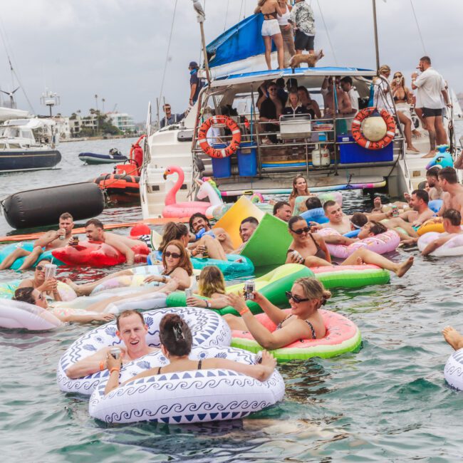 A large group of people relax on colorful inflatable floats in the water near a boat during a lively party. Some are swimming, while others sit on the boat. The scene is festive and crowded, with city buildings visible in the background.