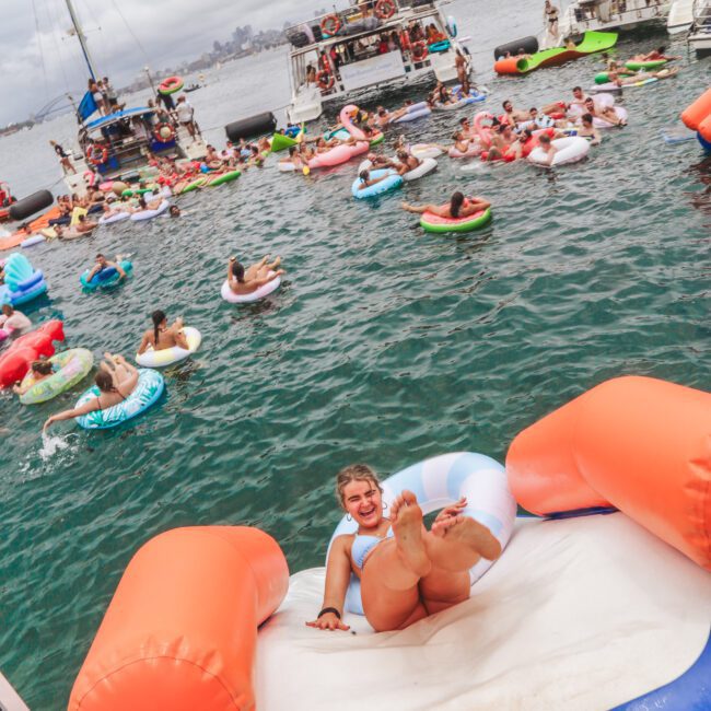 A woman laughs while lounging on a large inflatable float shaped like a chair in the water, surrounded by people on colorful inflatables near anchored boats under a cloudy sky.