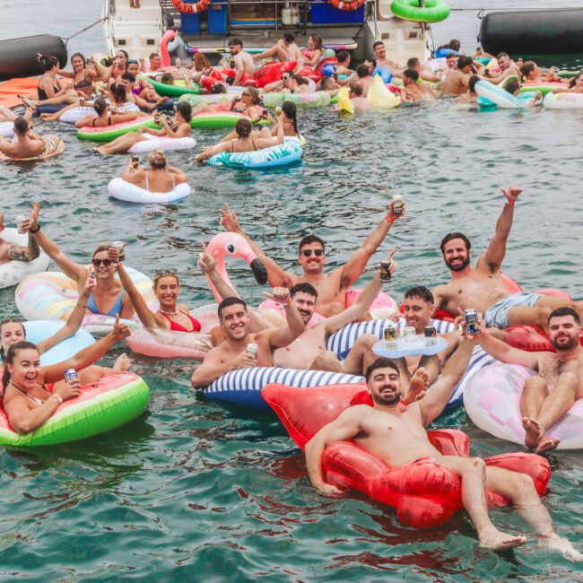 A large group of people relaxing on colorful inflatable pool floats in the water, smiling, laughing, and raising their drinks near a boat on a sunny day. The atmosphere is lively and festive.