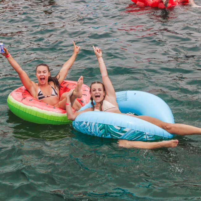 Two women in swimsuits float on colorful pool inflatables in a lake, smiling and raising their arms in excitement. One holds a drink can. The water around them is a deep green, and other floaters are in the background.