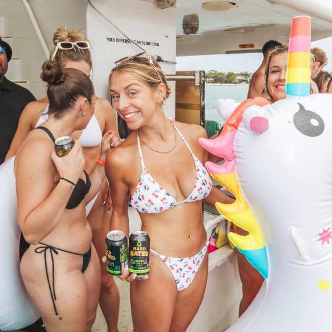 A smiling woman in a white bikini with colorful patterns holds two cans, standing beside a unicorn pool float at a lively boat party with other people in swimsuits.