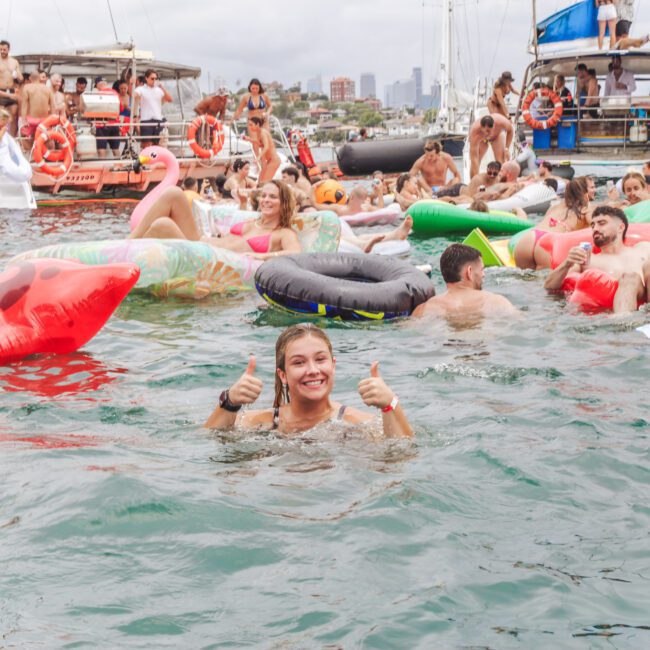 People enjoying a lively pool party on the water with colorful inflatable floats, boats in the background, and a smiling woman in the foreground giving two thumbs up while swimming.