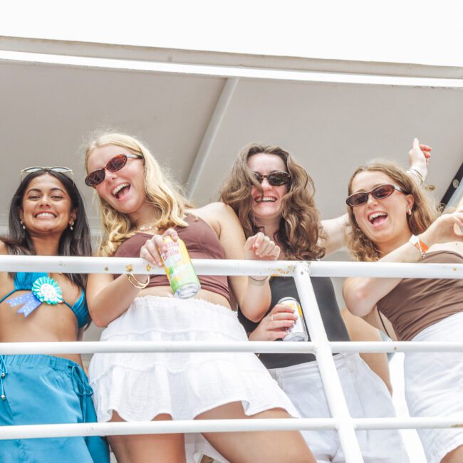 Four women smile and pose on a boat, holding drinks and wearing sunglasses and summer outfits. They appear to be celebrating and enjoying a sunny day outdoors.