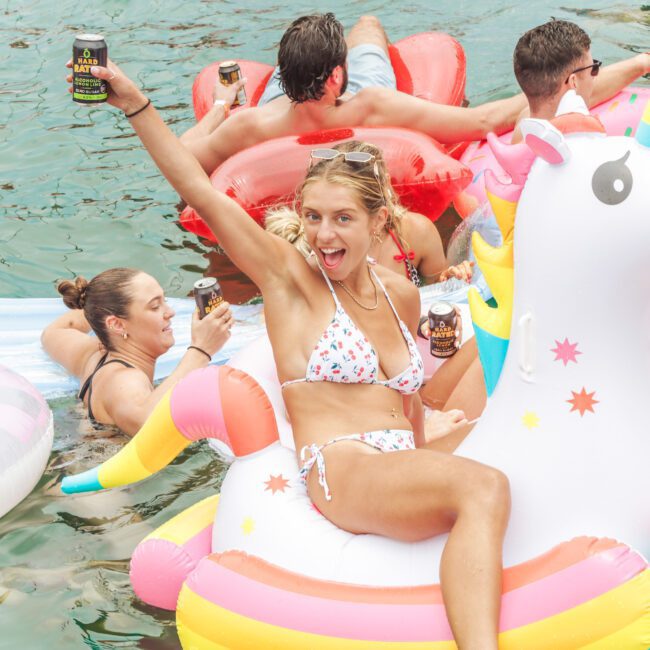 A smiling woman in a bikini raises a can while sitting on a unicorn pool float, surrounded by others on colorful floats, enjoying a sunny day on the water.