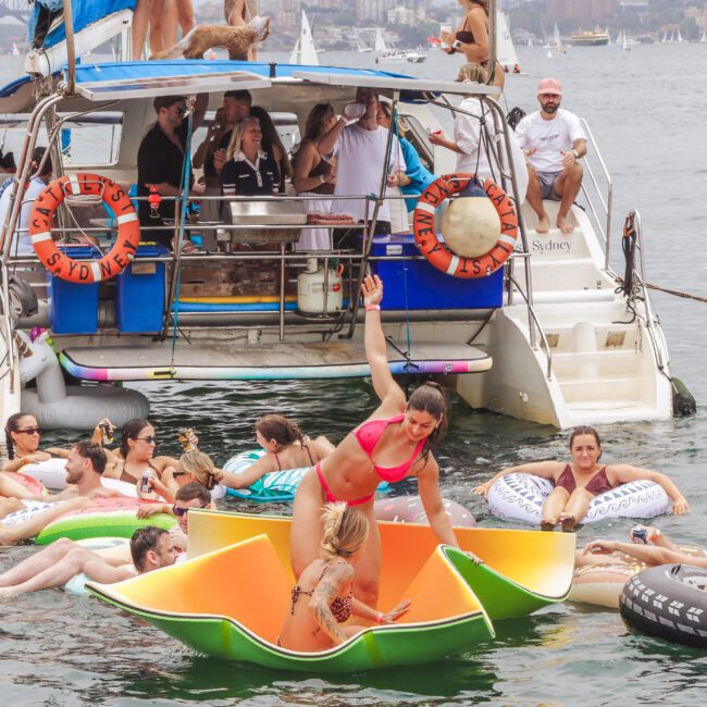 People enjoy a lively boat party on a sunny day, relaxing on colorful floaties in the water near a docked yacht, with many smiling, talking, and soaking up the festive atmosphere.
