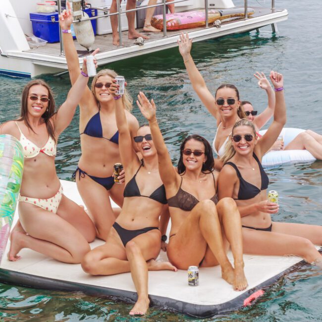 Six women in swimsuits sit on a floating mat in the water, smiling and raising their arms in celebration, with drinks in hand. A boat and another person on a float are visible in the background.