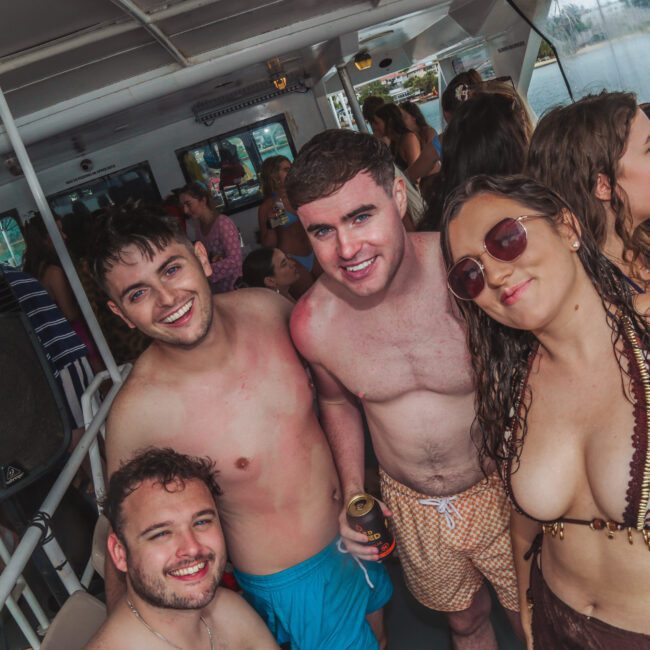 A group of young adults in swimsuits smile and pose together on a crowded boat party, surrounded by others enjoying drinks and socializing.
