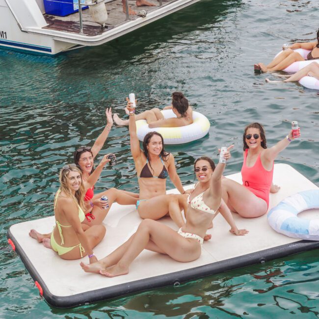 Six women in swimsuits sit and pose with drinks on a floating mat in the water, smiling and enjoying themselves. Other people on floaties and a boat are visible in the background. The scene is lively and summery.