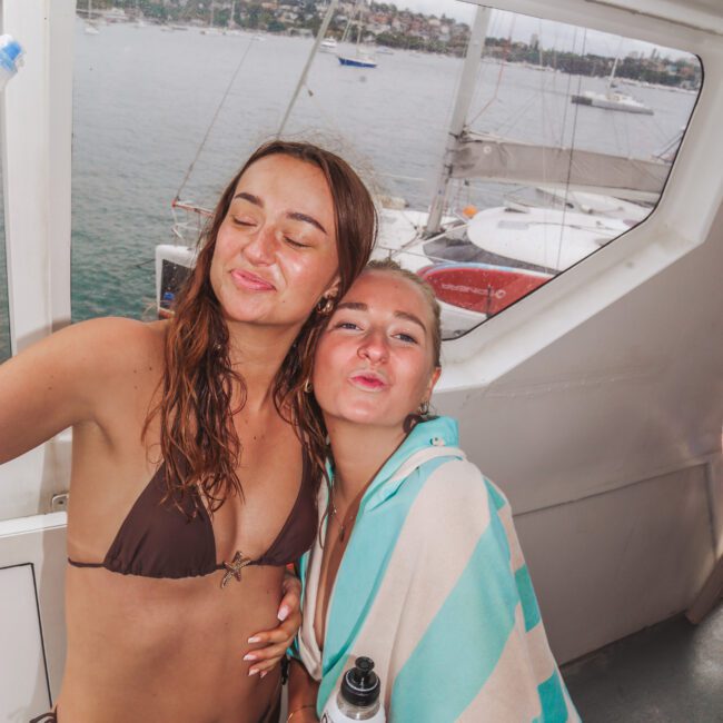 Two women in swimsuits pose playfully on a boat, one wrapped in a striped towel and holding a drink bottle. They pout for the camera, with water and anchored boats visible through the window behind them.
