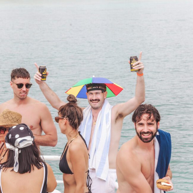 A group of people in swimsuits enjoy a boat party on the water; a man in the center wears a rainbow umbrella hat, holds drinks, and smiles with arms raised. Others chat, eat, and wear sunglasses and hats.