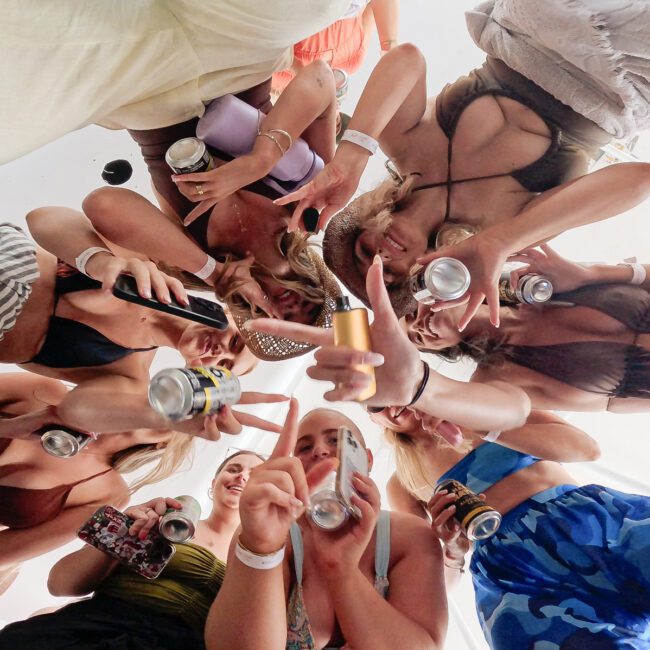 A group of women in swimsuits stand in a circle, holding drinks and cans, smiling and posing for a photo taken from below, creating a fun and festive atmosphere.