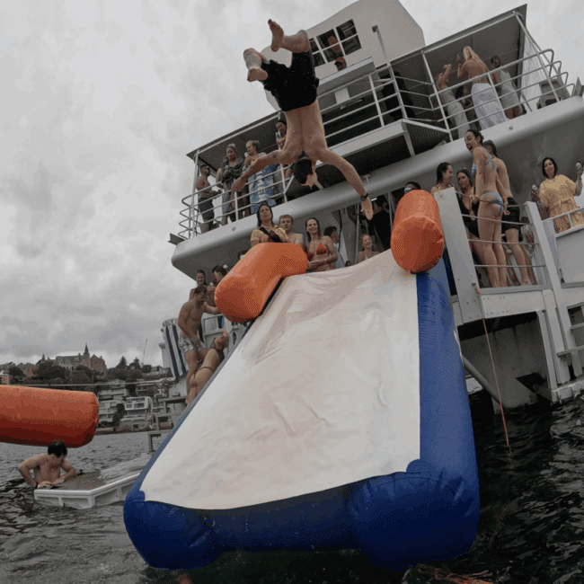 A person does a backflip off the end of an inflatable water slide attached to a boat, with a crowd of people watching and enjoying the scene on a cloudy day by the water.