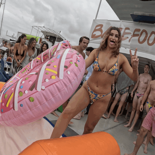 A smiling woman in a colorful bikini makes a peace sign while standing on an inflatable donut at a lively pool party with other people in swimwear. Boats and a "BAREFOOT" sign are visible in the background.
