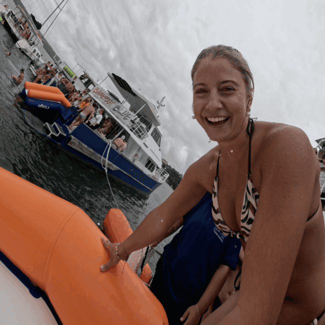 A smiling woman in a patterned bikini is holding onto an orange inflatable raft on the water, with a boat and several people in the background under a cloudy sky.