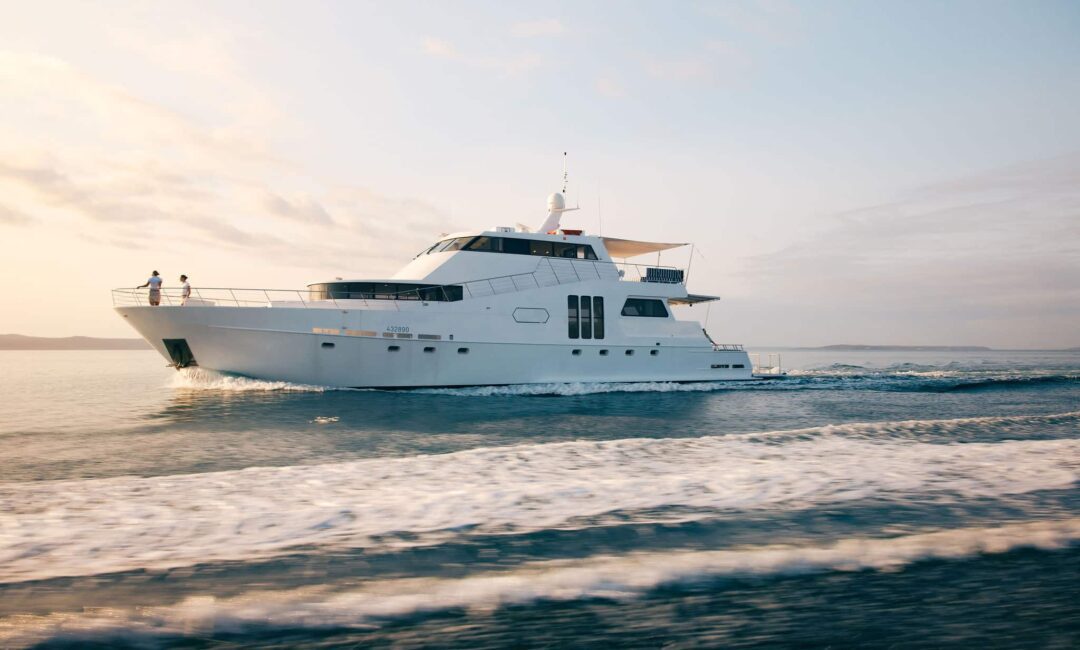 The YOT Viva glides across the water at sunset, leaving a gentle wake. Two people stand at the bow taking in the view, with a partly cloudy sky and a clear horizon stretching out ahead.