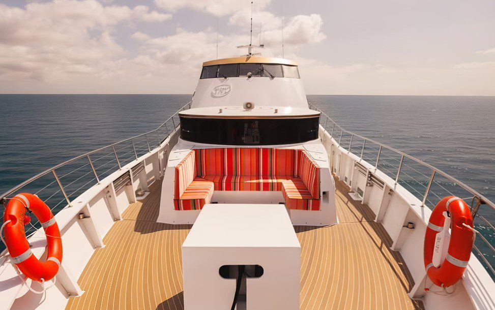 Enjoy the view from the YOT Viva deck with striped red and white seating, a white table, and two life rings on each side, overlooking calm ocean waters beneath a partly cloudy sky.