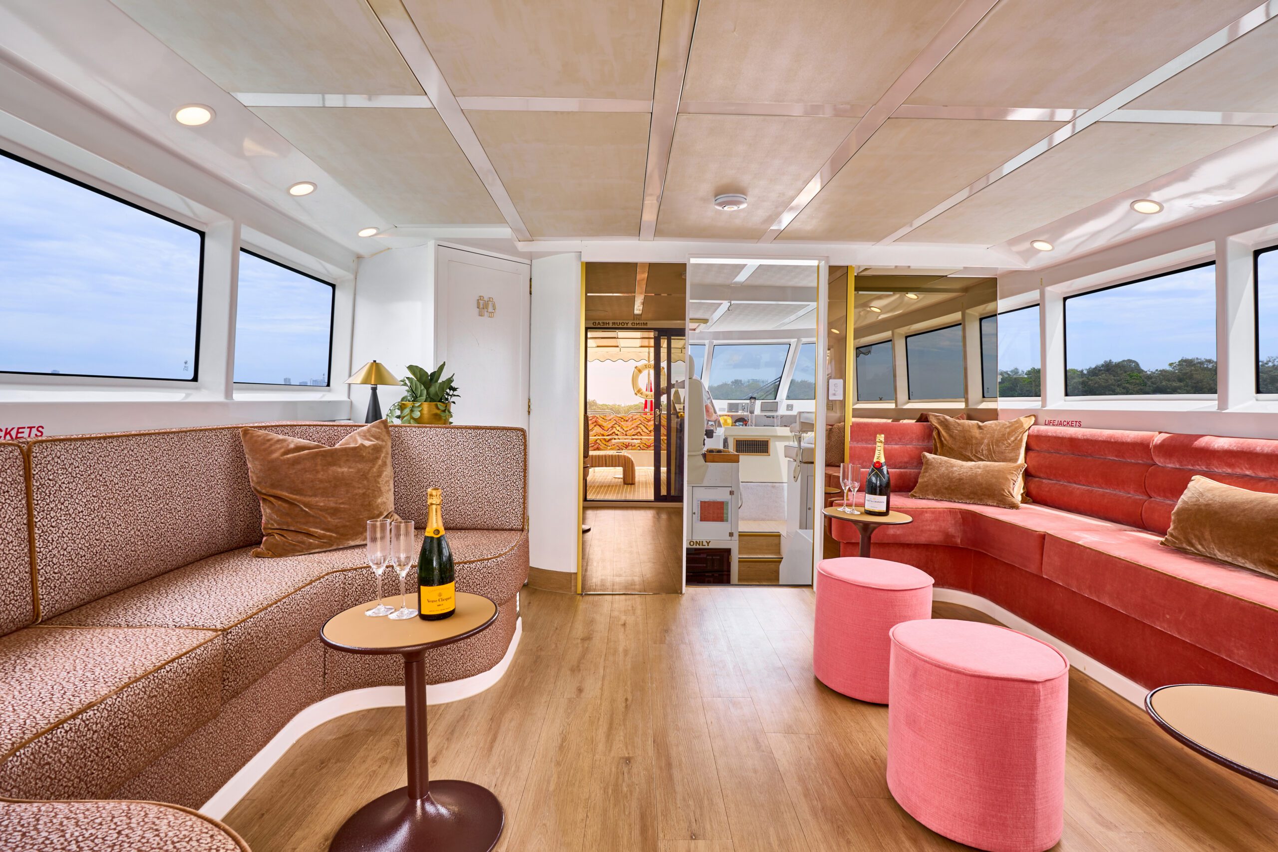 Modern boat interior with large windows, tan and red cushioned seating, small round tables with champagne and glasses, pink ottomans, and wood flooring. Natural light fills the space, creating a relaxed, elegant atmosphere.