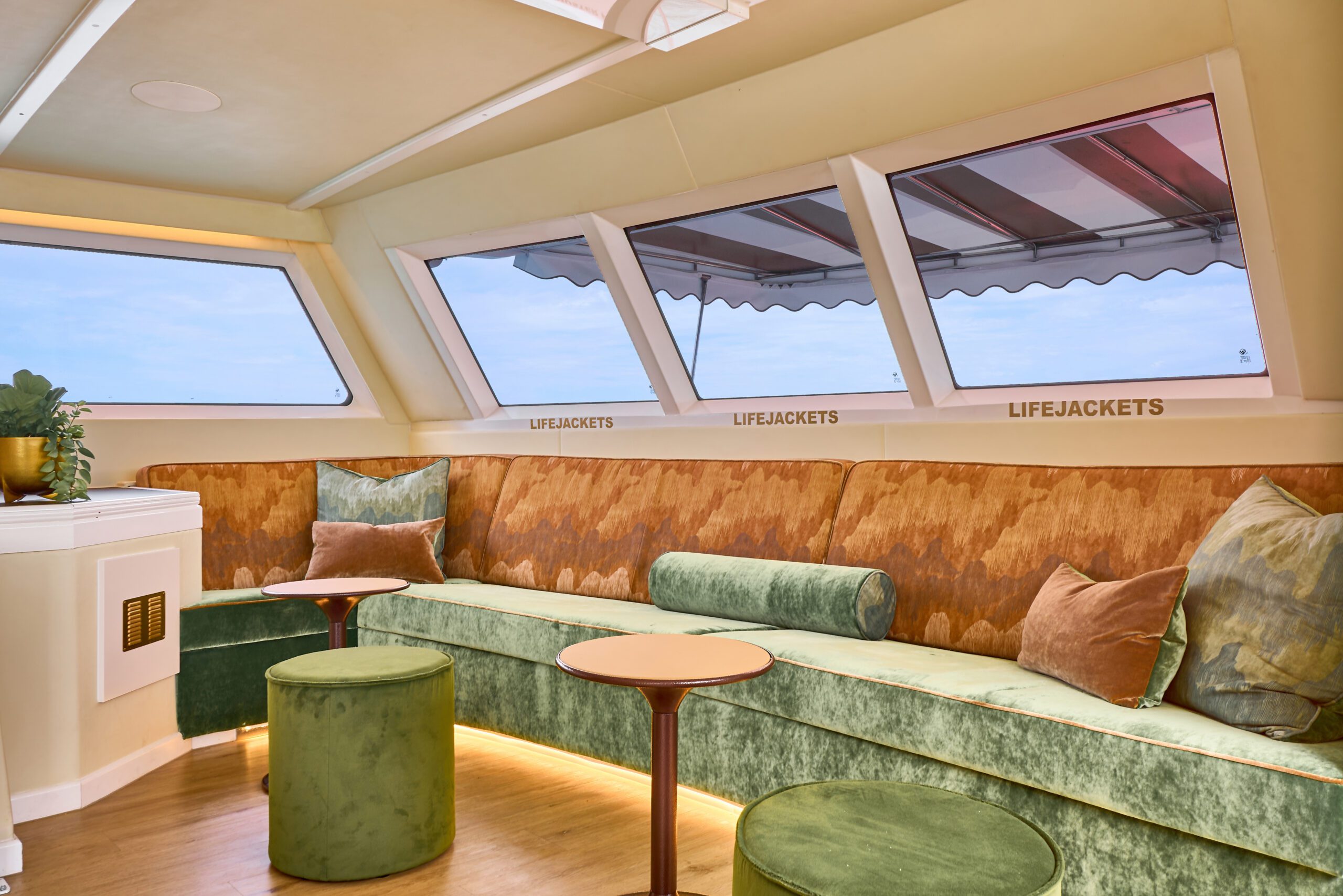 A modern boat lounge with green velvet seating, round tables, and large windows showing the sea. Cushions accent the seats, and "LIFEJACKETS" signs are visible under the windows.