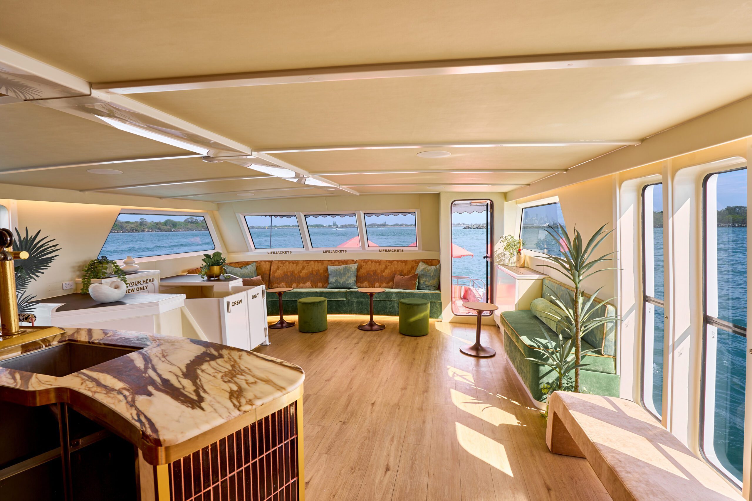 A bright, modern boat interior features green velvet seating, wooden floors, potted plants, a marble counter, large windows, and views of calm blue water and distant greenery.