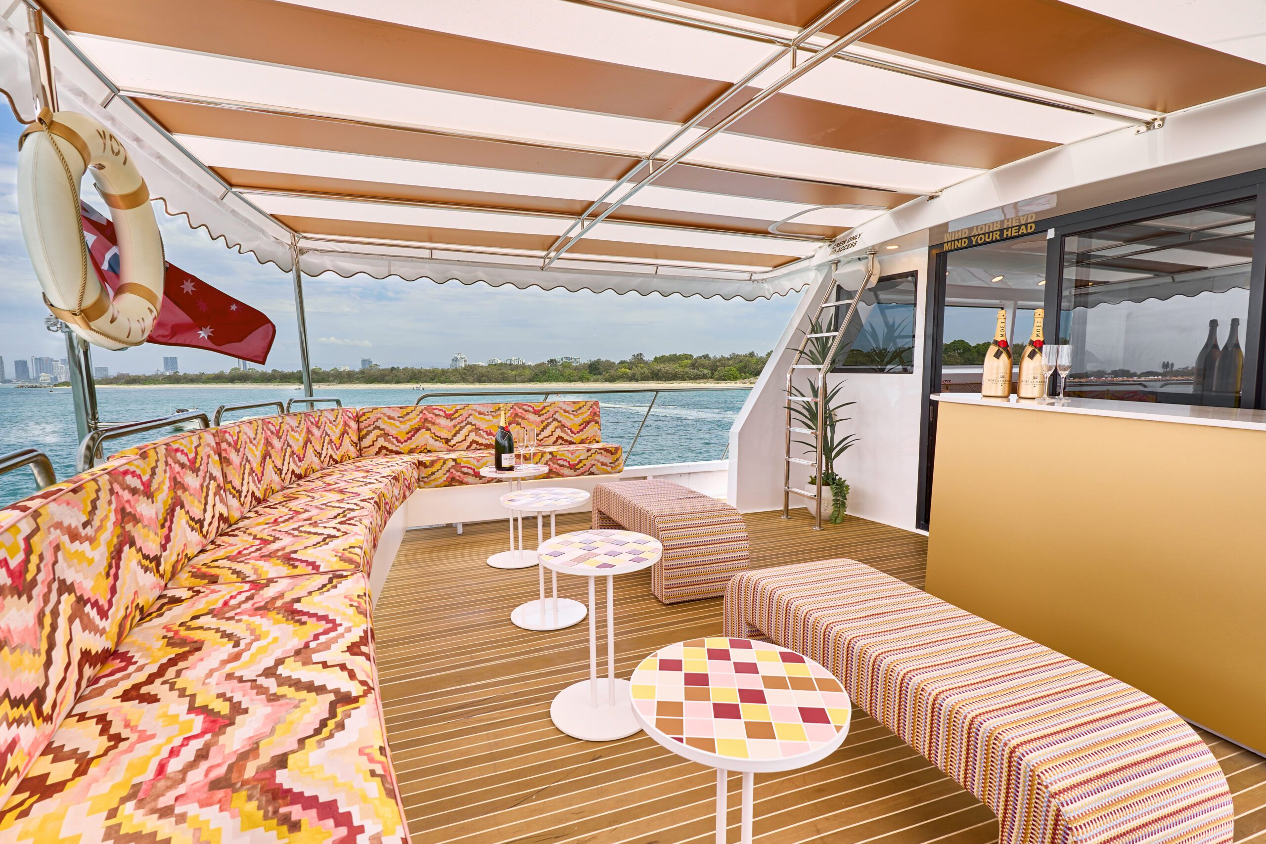 A luxury boat deck with colorful zigzag-patterned seating, small round and square tables, a bar with wine bottles, and a view of water and greenery in the background.