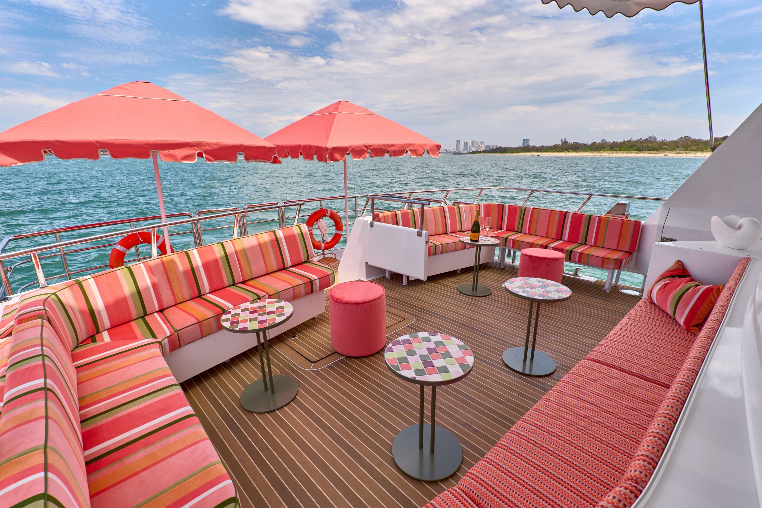 A yacht deck with colorful striped couches, pink ottomans, and small round tables under red umbrellas, overlooking blue ocean water and a distant shoreline under a partly cloudy sky.