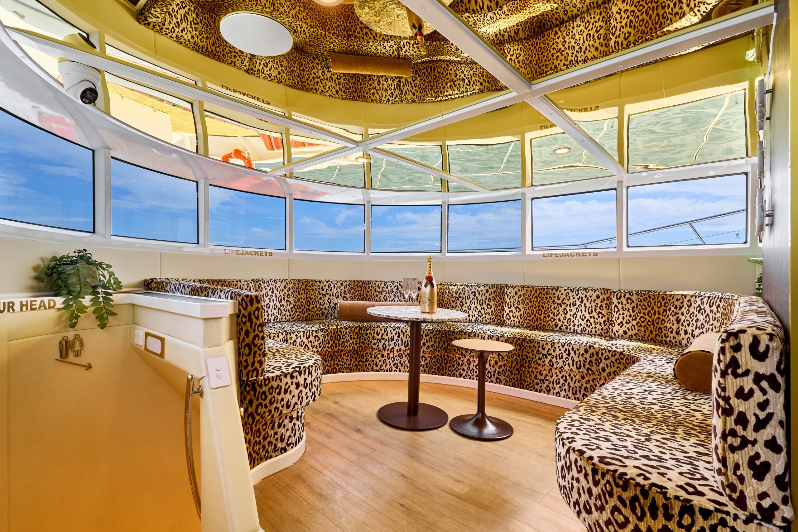 A room with curved leopard-print seating, two small round tables, a champagne bottle, large windows with views of the ocean and sky, mirrored ceiling panels, and a plant on a white counter.