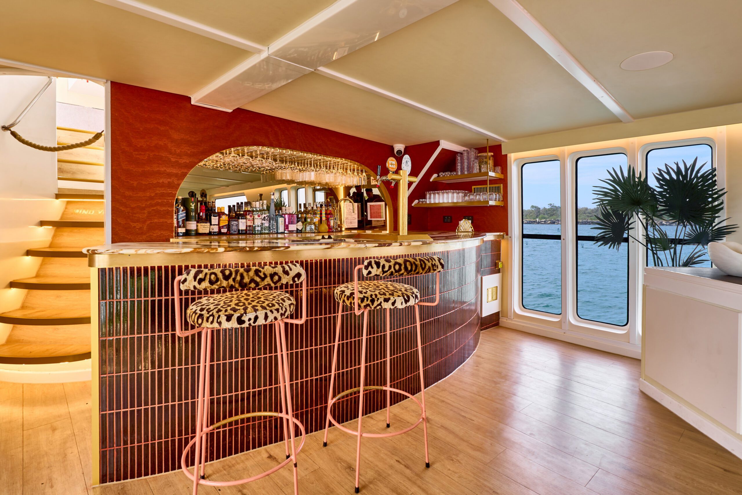 A stylish bar with a curved counter, two pink stools with leopard-print seats, various bottles and glasses, a mirrored back wall, wooden floor, and large windows overlooking water. Spiral stairs are visible on the left.