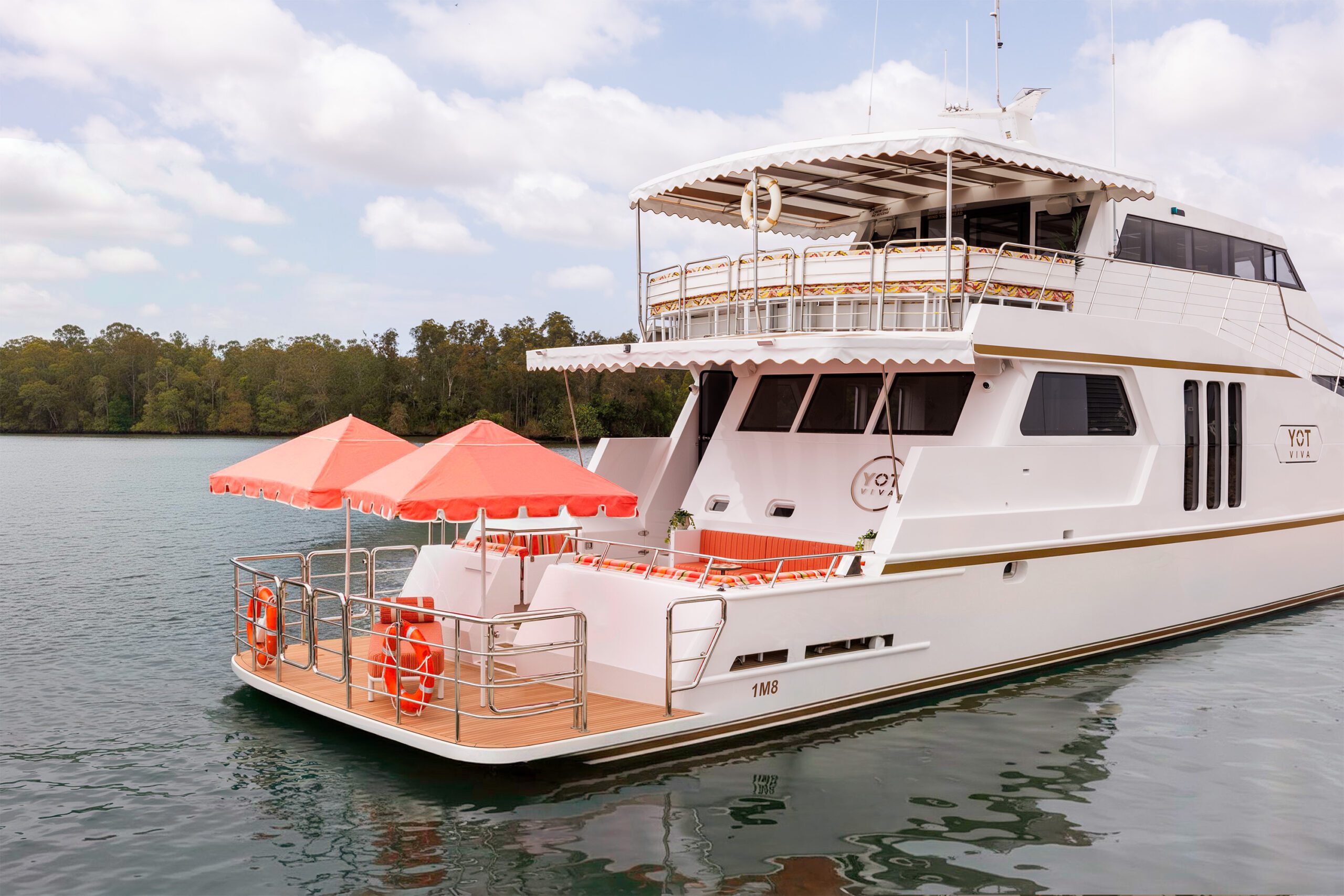 A white yacht with red-and-white umbrellas and orange life rings is docked on calm water near a tree-lined shore, under a partly cloudy sky.