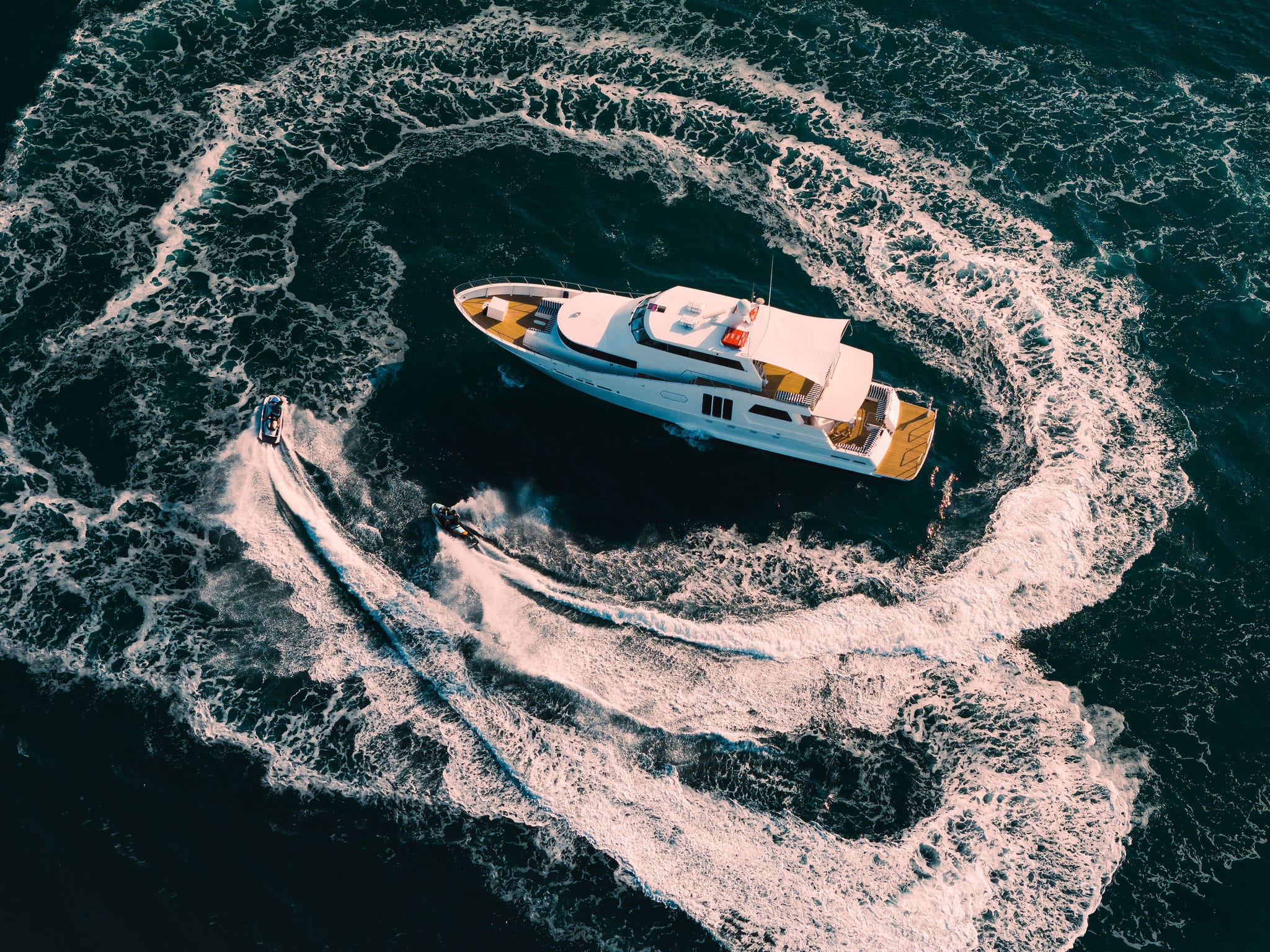 Aerial view of YOT Viva, a large white yacht, in deep blue water with two jet skis circling it and creating swirling white wakes.