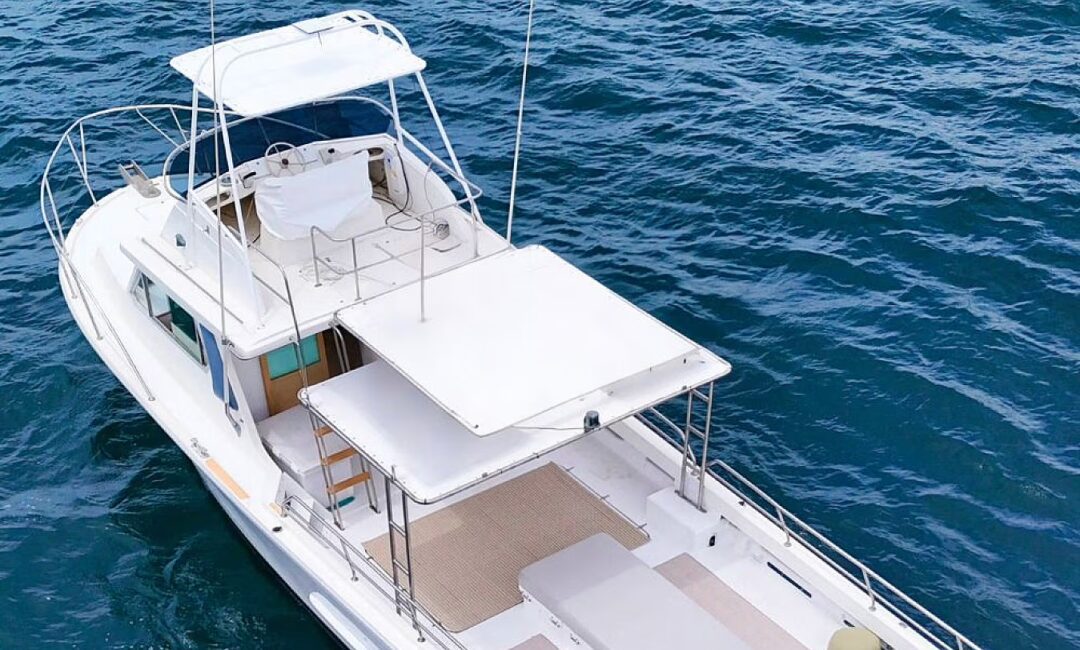Yachty, a white motor yacht with two covered decks, floats on calm blue water in an aerial view. The boat offers spacious seating and shaded areas, with no passengers visible aboard.