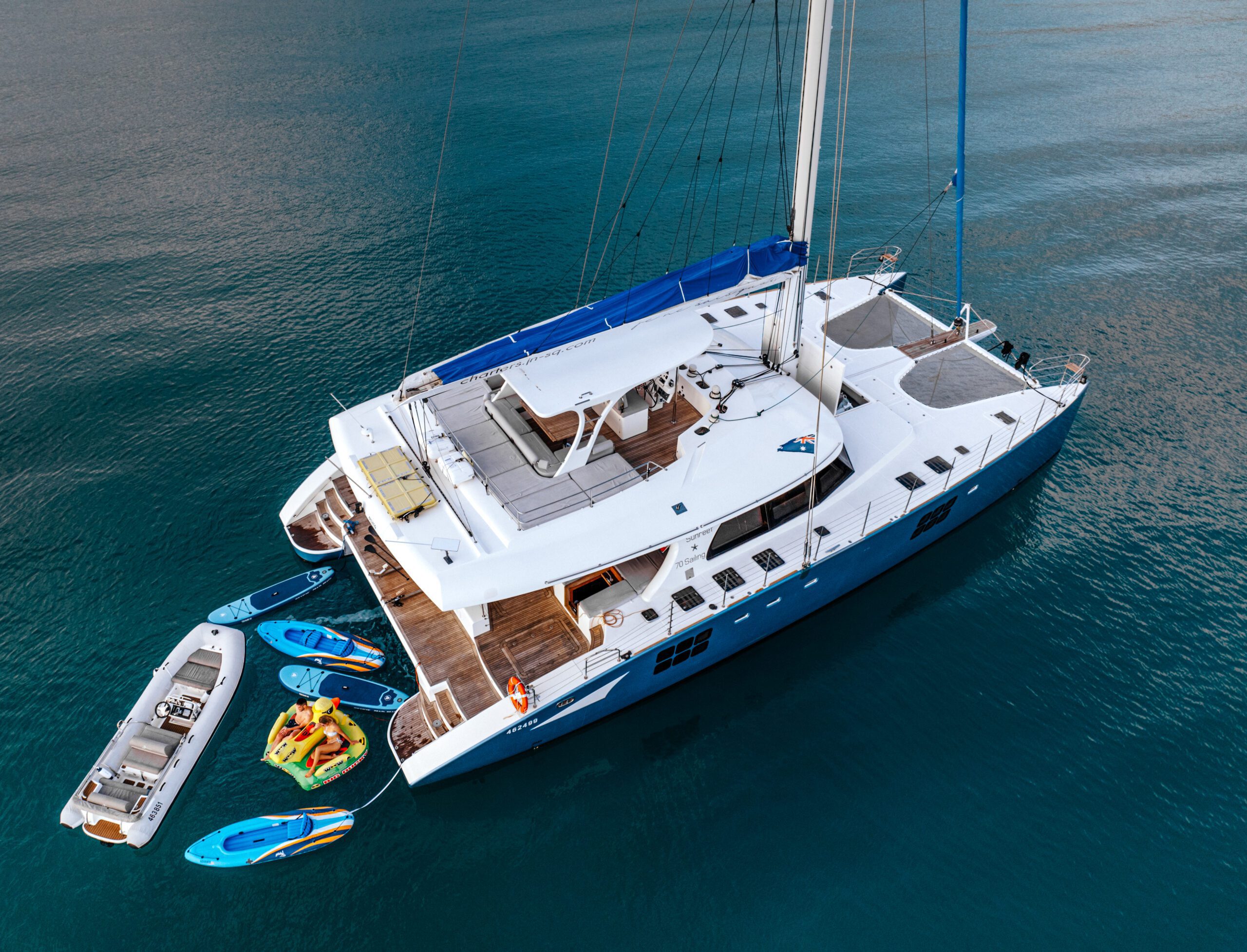 A large white catamaran yacht anchored in calm blue water, with a small inflatable boat, three paddleboards, and an inflatable water toy floating nearby. The yacht’s deck is spacious and well-equipped.