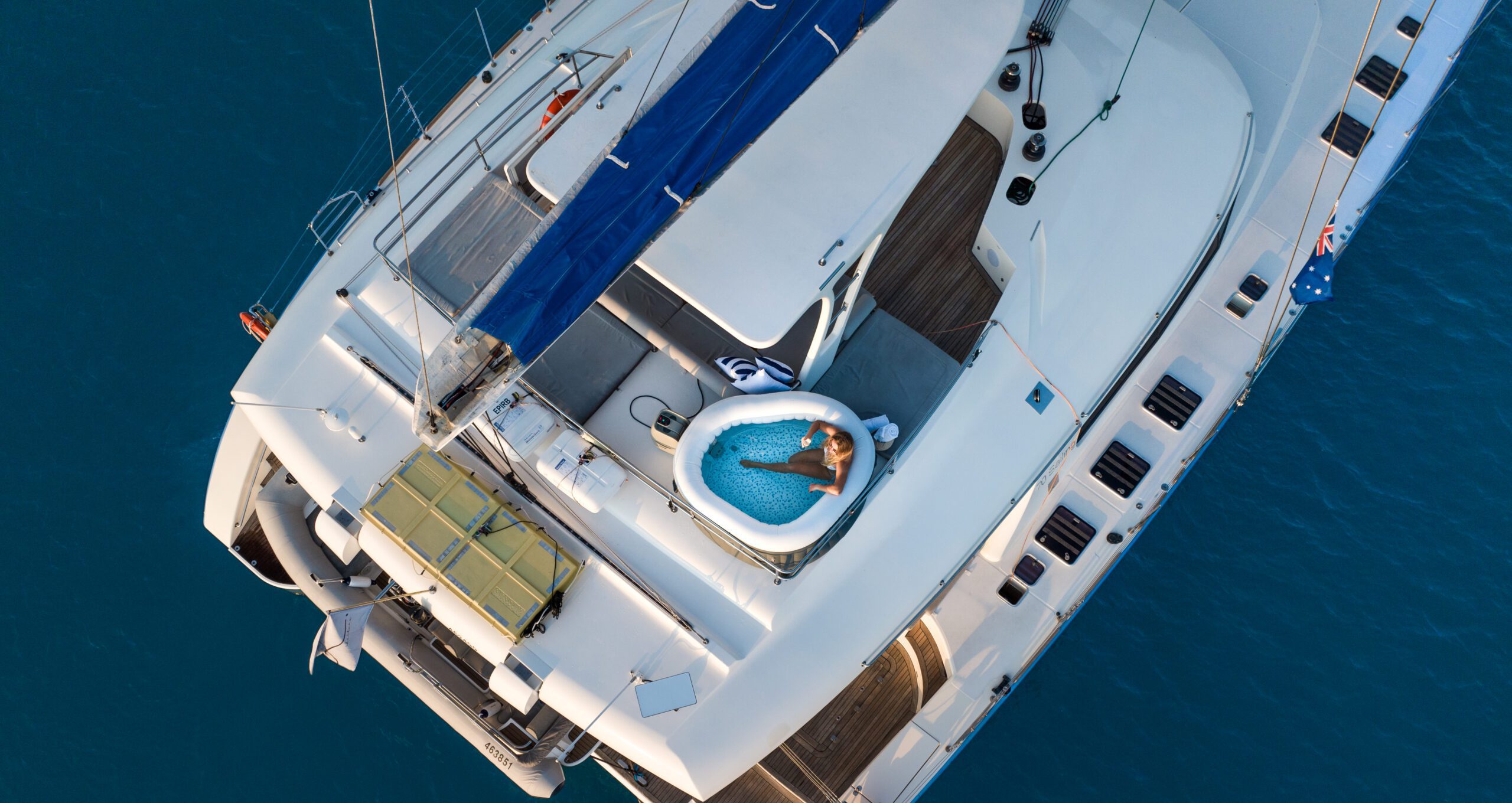 Aerial view of a yacht on blue water, with a woman relaxing in a small round hot tub on the deck, surrounded by white surfaces and blue sail covers.