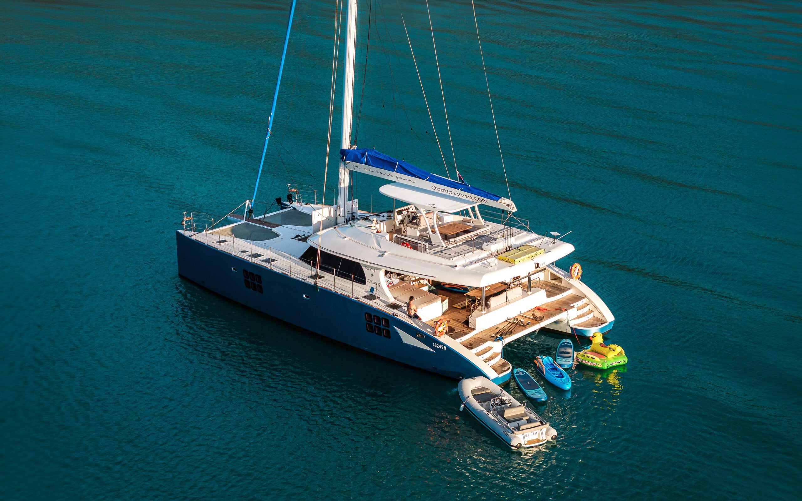 A large modern catamaran yacht floats on calm blue water, with several kayaks, paddleboards, and an inflatable raft tied behind it, and a small motorboat alongside.