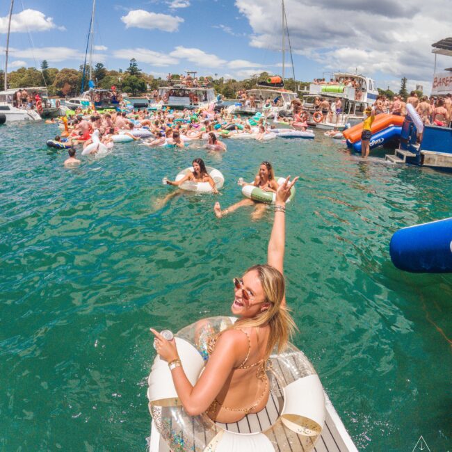 A woman in sunglasses and a floaty sits on a dock, smiling and raising her arm toward a lively group of people in pool floats enjoying a sunny party on the water, with boats and blue skies in the background.
