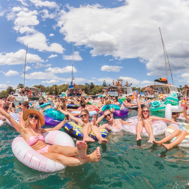 A large group of people relax on colorful inflatable floats in a lively lake party, smiling under a sunny sky with scattered clouds, surrounded by boats and vibrant summer energy.