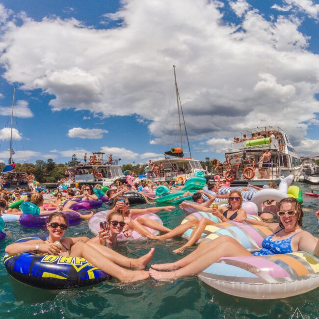 A group of people relax on colorful inflatable floats in the water at a lively boat party, with boats and more partygoers in the background under a partly cloudy sky.