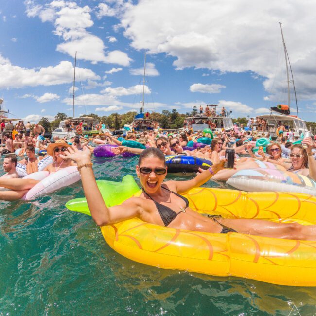A smiling woman in sunglasses lounges on a pineapple-shaped float in a crowded lake full of people on inflatables, with boats and blue sky in the background. The scene is lively and festive.