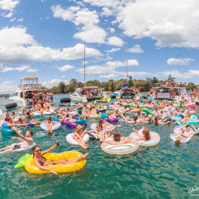 A large group of people on colorful inflatable floats relax in a lake near several anchored boats under a partly cloudy sky, enjoying a lively outdoor gathering on the water.