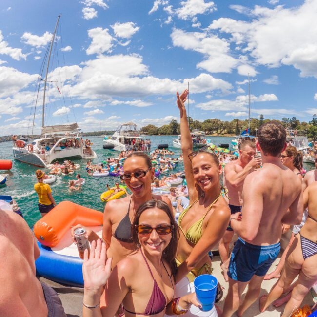 A group of people in swimsuits smile and wave at the camera at a lively boat party on the water, surrounded by other boats, inflatables, and a crowd enjoying a sunny day.