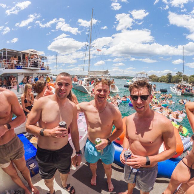 Three young men in swimwear stand on a boat holding drinks, smiling at the camera. Behind them, a large group of people are swimming and relaxing on inflatables in the water, with boats and a blue sky visible.