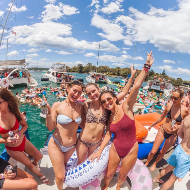 Three women in swimsuits pose and smile on a crowded boat party, surrounded by people, colorful floaties, and drinks, with boats and clear turquoise water under a bright blue sky with clouds.