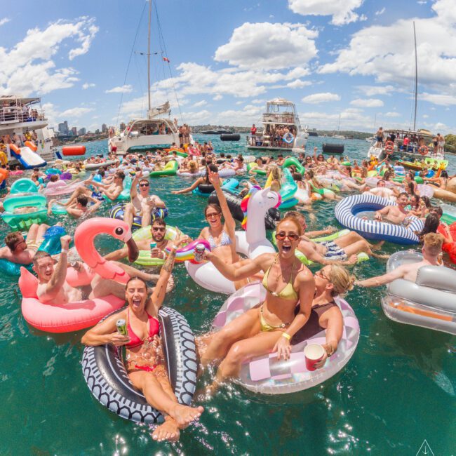 A large group of people relax and have fun on colorful inflatable floats in the water near anchored boats on a sunny day, with blue skies and fluffy clouds overhead.
