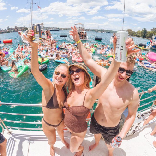 Three young adults in swimsuits smile and raise drinks on a boat, with a lively crowd on colorful floats and inflatables in the water behind them, under a bright, sunny sky.