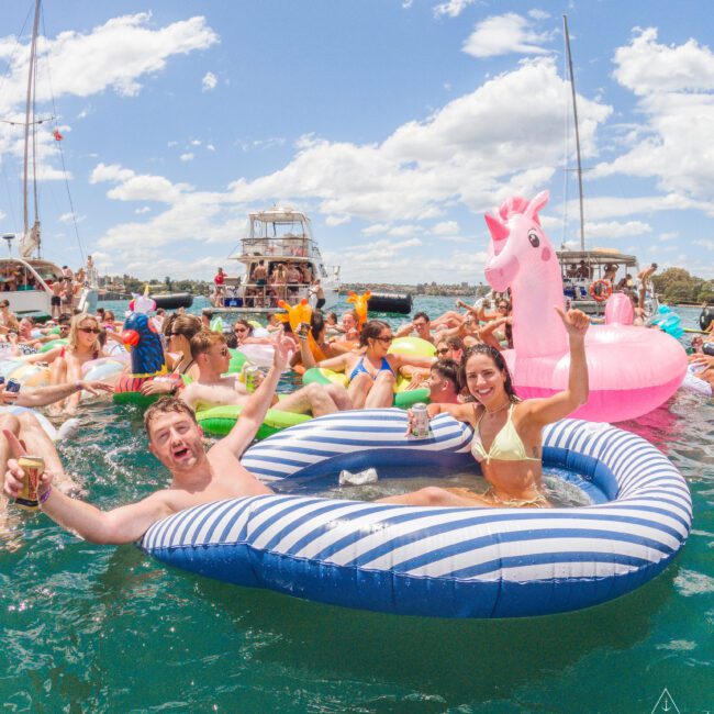A group of people enjoy a lively pool party on the water, floating on colorful inflatables, including a striped raft and a pink unicorn, under a sunny sky with boats in the background.