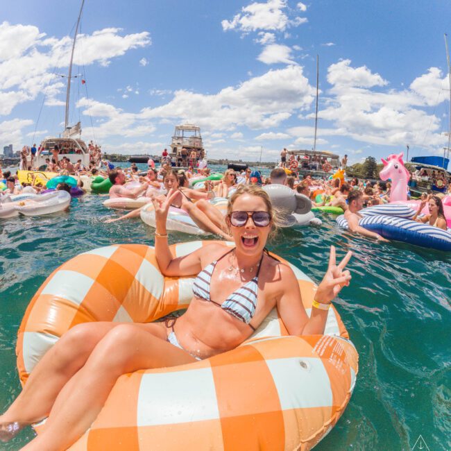 A woman in a bikini smiles and flashes peace signs while relaxing on an orange checkered float in a busy, sunny water scene with many people on inflatable rafts and boats in the background.