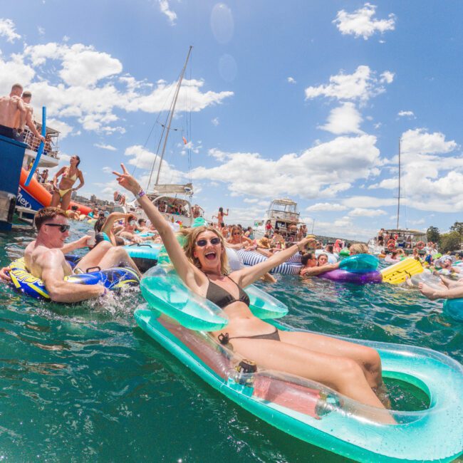 A group of people relax and have fun on colorful inflatables in the water near boats. A smiling woman in sunglasses and a bikini floats in the foreground, raising her arm and making a peace sign under a sunny sky.