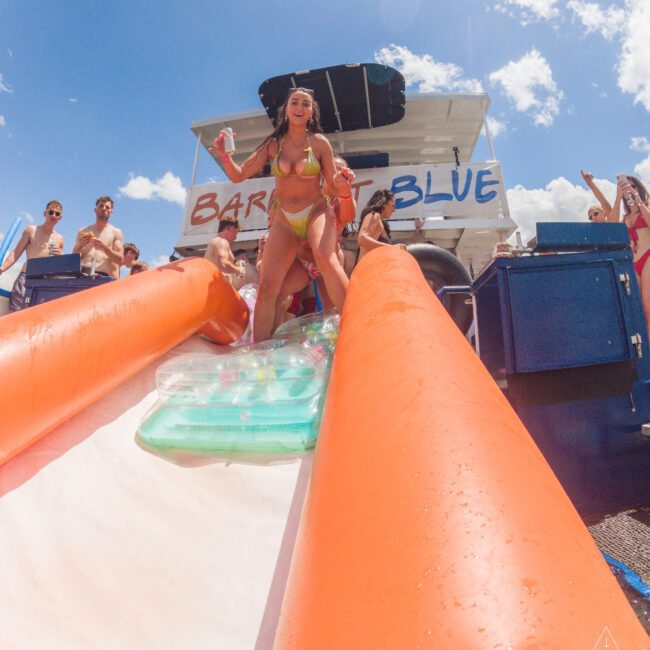 A woman in a bikini holds a drink and smiles while standing at the top of an inflatable water slide on a boat named "Bar Blue," surrounded by people enjoying a sunny day.