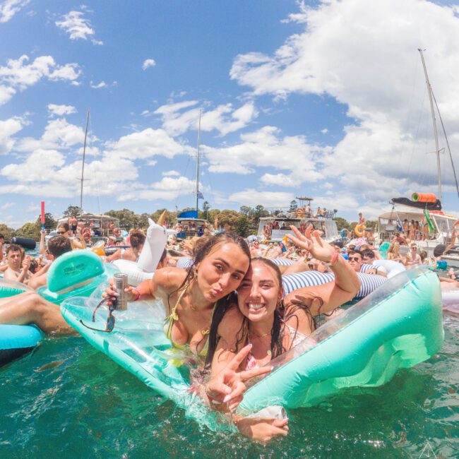 Two women smiling and posing on a green pool float in the water, surrounded by a crowd of people on floats and boats at a lively summer party under a blue sky with clouds.